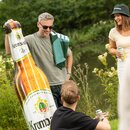 Mann mit Sonnenbrille hält eine aufblasbare Krombacher-Pils-Flasche, umgeben von zwei weiteren Personen mit Getränkedosen in der Natur