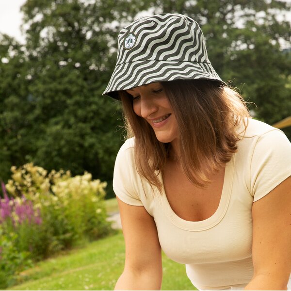 Frau mit Krombacher Wappen und wellenförmig gemustertem Bucket Hat und weißem T-Shirt lächelnd im Garten