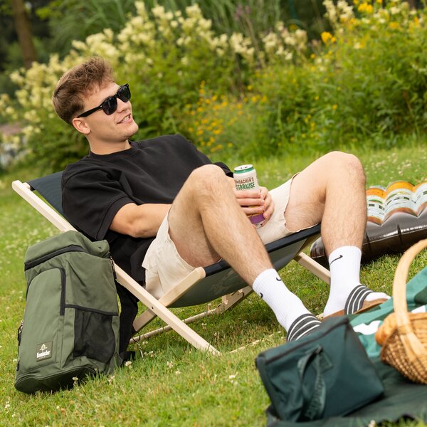 Junger Mann mit Sonnenbrille und schwarzem T-Shirt sitzt entspannt in einem Liegestuhl auf einer Wiese und hält eine Getränkedose