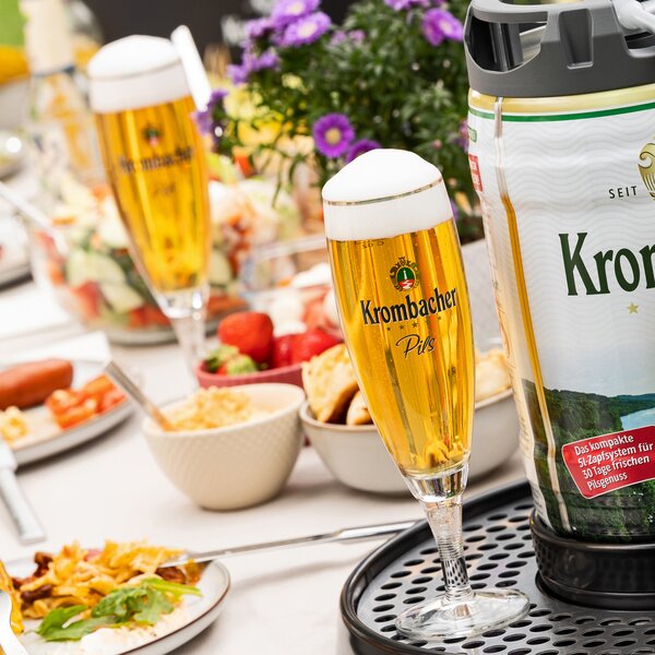 Zwei gefüllte Krombacher Pils-Gläser mit Schaum auf einem gedeckten Tisch mit Snacks und Blumen im Hintergrund