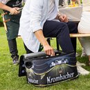 Eine Person greift in eine offene Krombacher-Kühltasche, die auf einer grünen Wiese vor einer Bierbank steht, während im Hintergrund gegrillt wird.