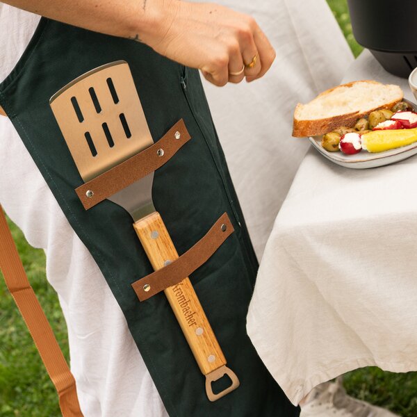 Grillzubehör mit Holzgriff in Lederschlaufen an schwarzer Schürze befestigt neben gedecktem Gartentisch mit Brot und Gemüse