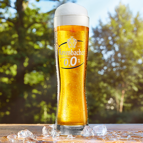 Krombacher 0,0% in einem hohen, schlanken Glas, umgeben von Eiswürfeln vor unscharfem Waldhintergrund.
