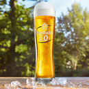 Krombacher 0,0% in einem hohen, schlanken Glas, umgeben von Eiswürfeln vor unscharfem Waldhintergrund.