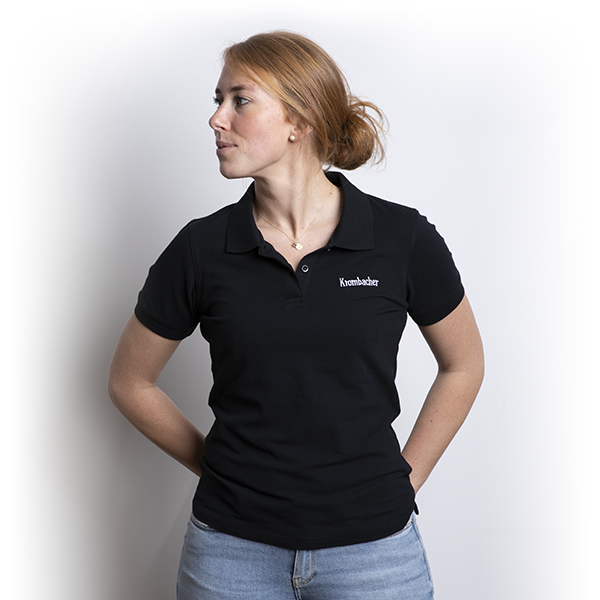 Frau trägt schwarzes Krombacher Damen Poloshirt mit kurzem Arm und weißem Logo auf der Brust, steht mit Händen in den Hüften.