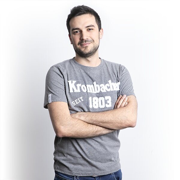 Mann mit verschränkten Armen trägt graues T-Shirt mit weißem Schriftzug 'Krombacher seit 1803'.