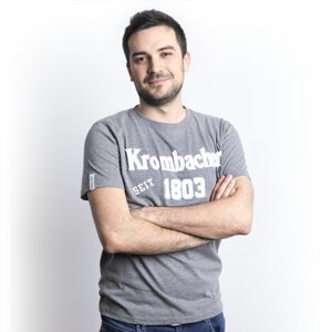 Mann mit verschränkten Armen trägt graues T-Shirt mit weißem Schriftzug 'Krombacher seit 1803'.