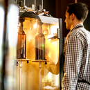 Mann in kariertem Hemd steht vor einer Abfüllmaschine mit zwei stehenden Glasflaschen im Inneren.