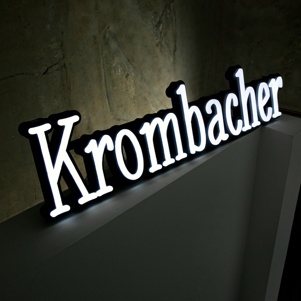 Strahlender Leuchtschriftzug von Krombacher in dunkler Umgebung vor Betonwand.