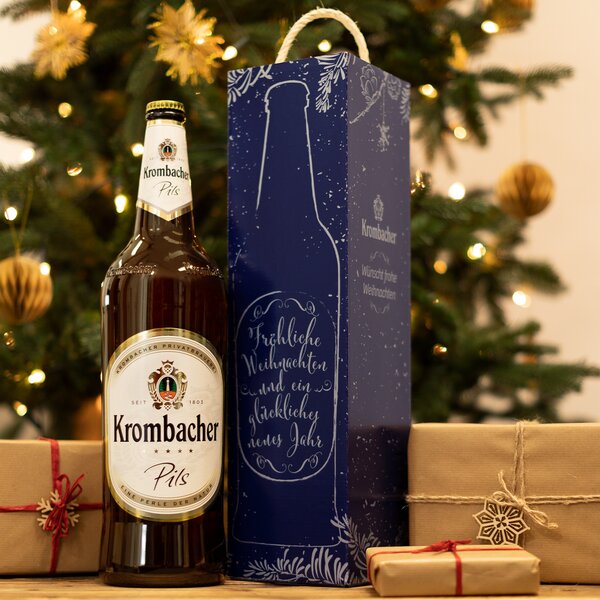 3-Liter Magnumflasche Krombacher Pils neben blauer Geschenkbox mit Weihnachtsgruß vor geschmücktem Weihnachtsbaum und verpackten Geschenken.